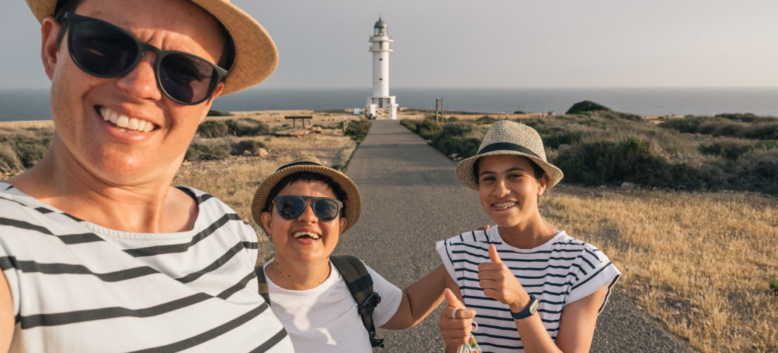 Drei Frauen machen ein Selfie auf einem sonnigen Küstenweg, im Hintergrund ein weißer Leuchtturm am Ende des Weges. Alle tragen Hüte, Sonnenbrillen und haben sommerliche Kleidung an. Die Stimmung ist fröhlich und ausgelassen, eine der Frauen zeigt einen D