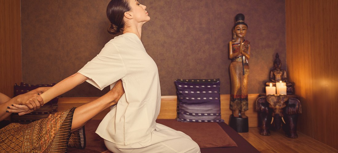 Frau erhält eine traditionelle Thai-Massage in einem stilvoll eingerichteten Raum mit Holzverkleidung. Die Masseurin nutzt Arme und Beine für eine Dehnungstechnik. Im Hintergrund stehen asiatische Dekofiguren und brennende Kerzen