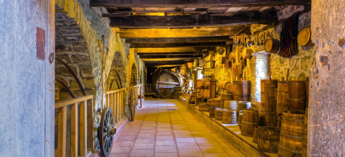 Innenraum eines alten Weinkellers mit Steinwänden und Holzbalken an der Decke. Entlang der Wände stehen zahlreiche hölzerne Weinfässer und antike Geräte zur Weinherstellung. Die Beleuchtung ist warm und schafft eine rustikale Atmosphäre. Wagenräder und We