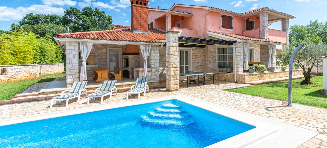Großes Ferienhaus mit rosa Fassade, Natursteinmauern und rotem Ziegeldach. Im Vordergrund befindet sich ein Pool mit Liegestühlen. Der Garten ist gepflegt mit Rasen, Olivenbaum und überdachter Terrasse mit Grill.