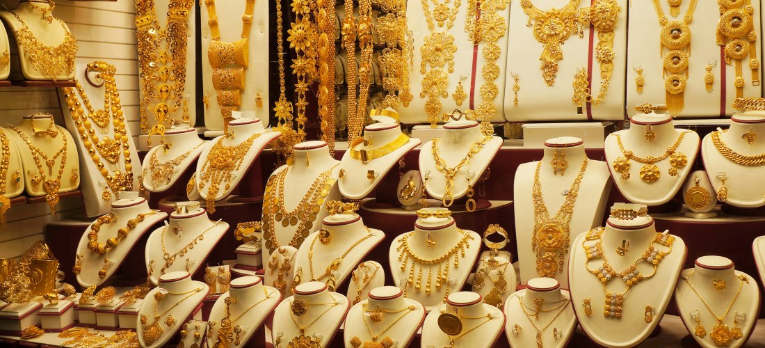 Auslage in einem Schaufenster des Gold Souk in Dubai. Zu sehen sind zahlreiche goldene Schmuckstücke wie Ketten, Ringe, Armbänder und Ohrringe, kunstvoll arrangiert auf weißen Büsten. Die Goldstücke funkeln im warmen Licht und zeigen orientalische Designs