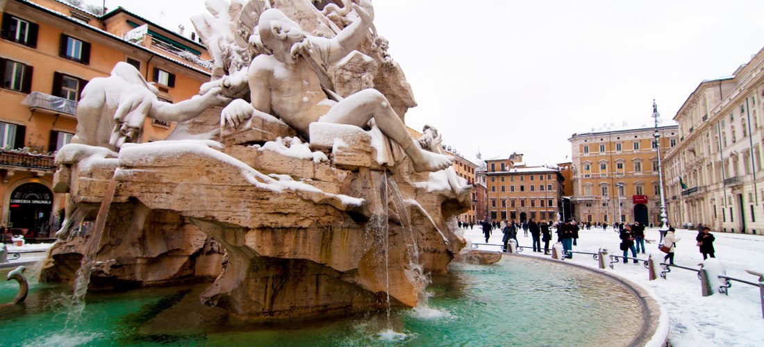 Verschneiter Vierströmebrunnen auf der Piazza Navona in Rom. Die barocke Brunnenfigur ist mit einer Schneeschicht bedeckt, Wasser fließt weiterhin in das grünlich schimmernde Becken. Menschen spazieren durch das winterliche Stadtzentrum.