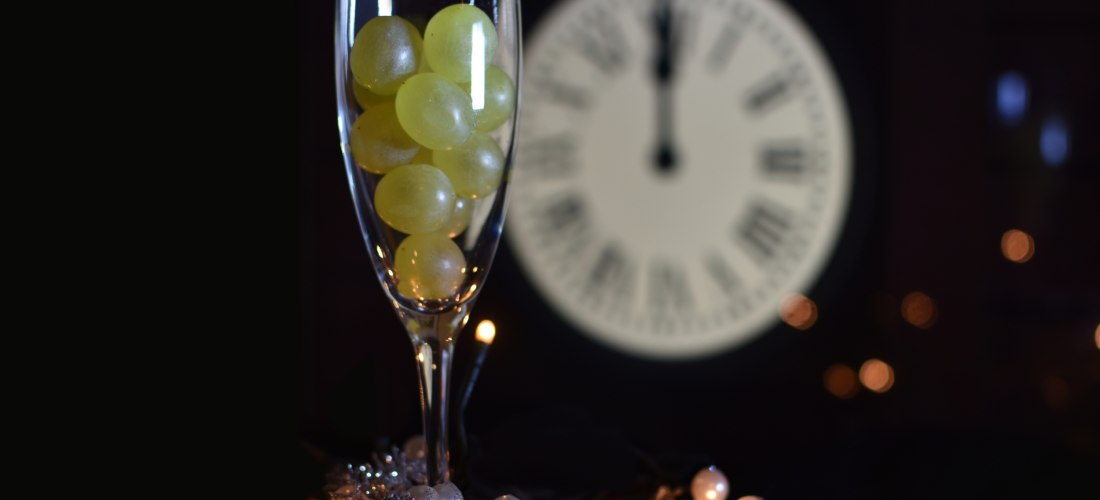 Ein Sektglas gefüllt mit zwölf grünen Trauben, im Hintergrund eine Uhr, die Mitternacht anzeigt – symbolisch für den spanischen Silvesterbrauch.