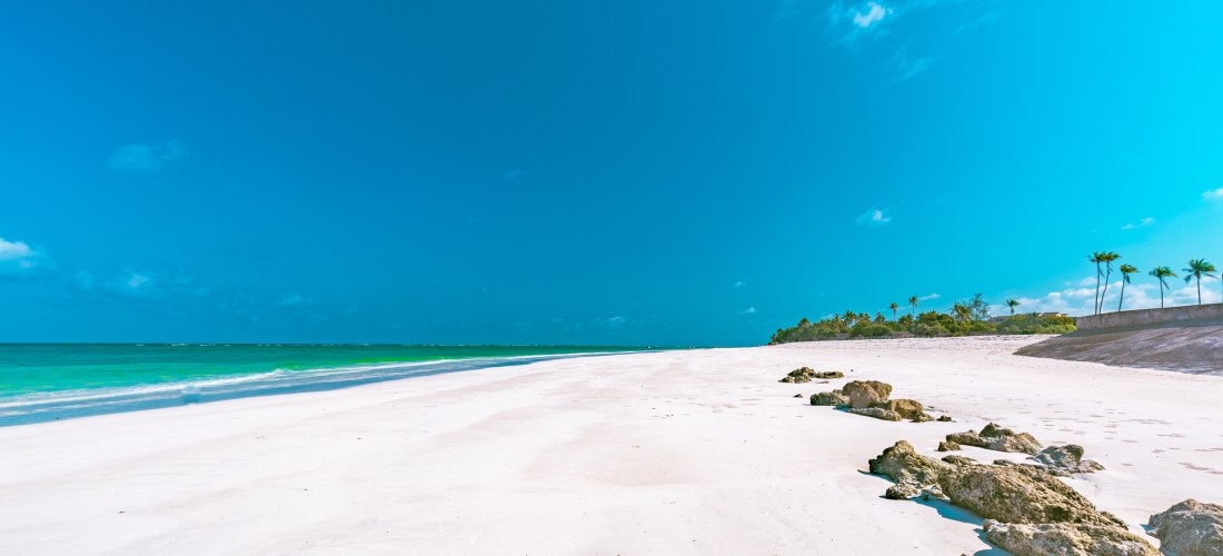 Ein weiter, menschenleerer Sandstrand mit strahlend weißem Sand und türkisblauem Meer unter wolkenlosem Himmel. Auf der rechten Seite Palmen und einige Felsen am Strand, im Hintergrund tropische Vegetation