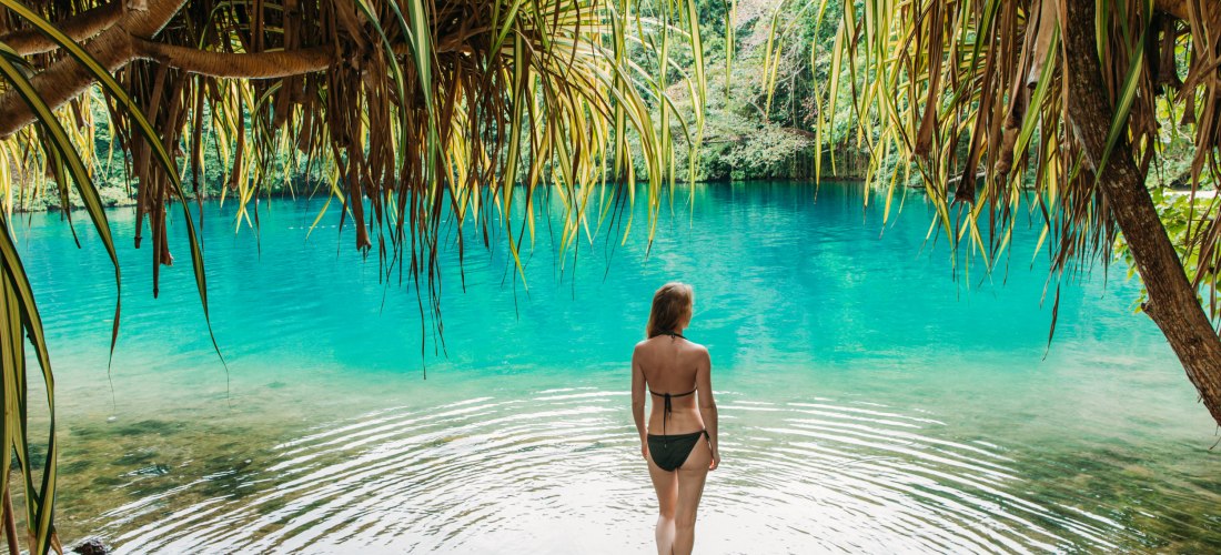 Frau im Bikini steht am Ufer einer türkisfarbenen Lagune, umgeben von tropischer Vegetation – idyllischer Naturpool inmitten des Dschungels.