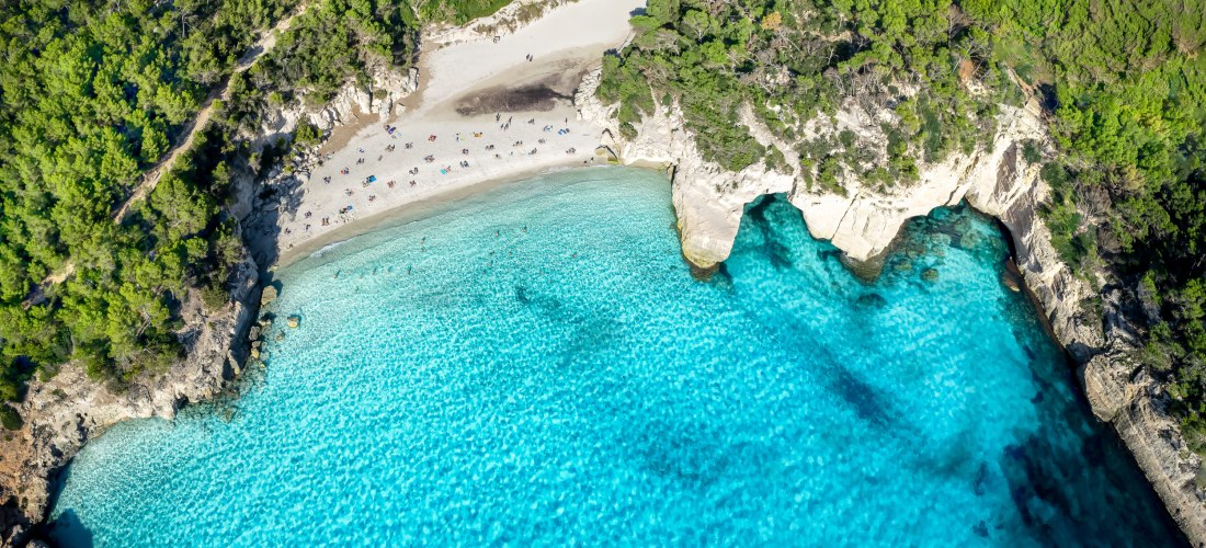 Luftaufnahme Cala Mitjana und Mitjaneta auf Menorca: feiner Sandstrand, umgeben von grünen Pinienwäldern, mit türkisblauem Wasser, in dem einige Badegäste schwimmen.