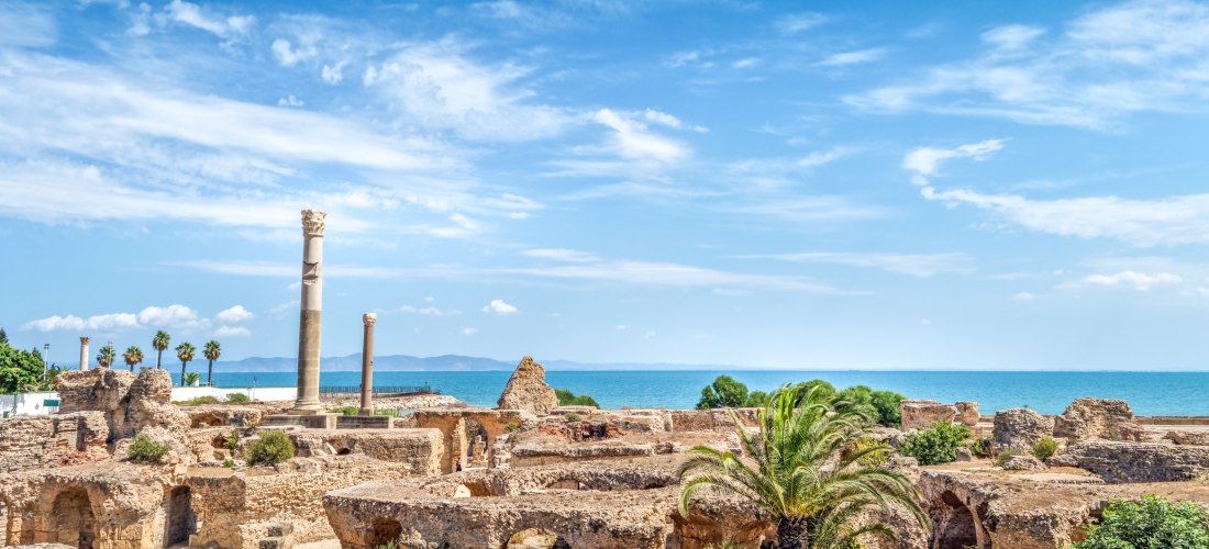 Antike Ruinen einer römischen Stadt nahe am Meer, mit einzelnen stehenden Säulen, Palmen und Überresten von Mauern. Der Himmel ist blau mit leichten Wolken, und das Meer erstreckt sich bis zum Horizont.