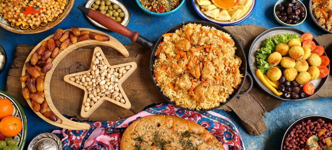 Bunt gedeckter Tisch mit traditionellen orientalischen Speisen: Reispfanne mit Hähnchen, Falafel, Oliven, Datteln, Fladenbrot, Kichererbsensalat, süße Gebäckstücke und Nüsse in einem Holztablett in Form von Halbmond und Stern