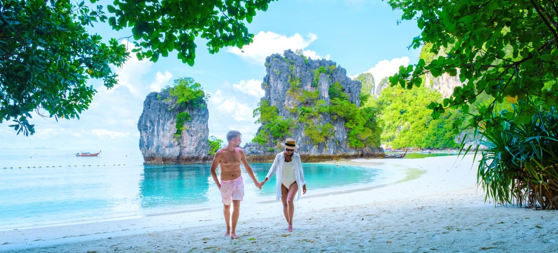 Das Bild zeigt ein Paar, das barfuß Hand in Hand an einem tropischen Sandstrand entlangspaziert. Im Hintergrund liegt kristallklares, türkisfarbenes Wasser, das sanft an den Strand schwappt. Die Szene wird eingerahmt von dichtem, grünem Blattwerk und mark
