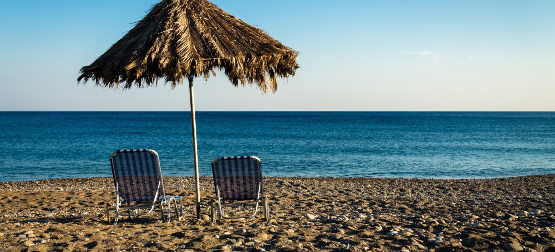 Zwei Liegestühle unter einem Strohdach am ruhigen Kiesstrand auf Kreta mit Blick aufs Meer