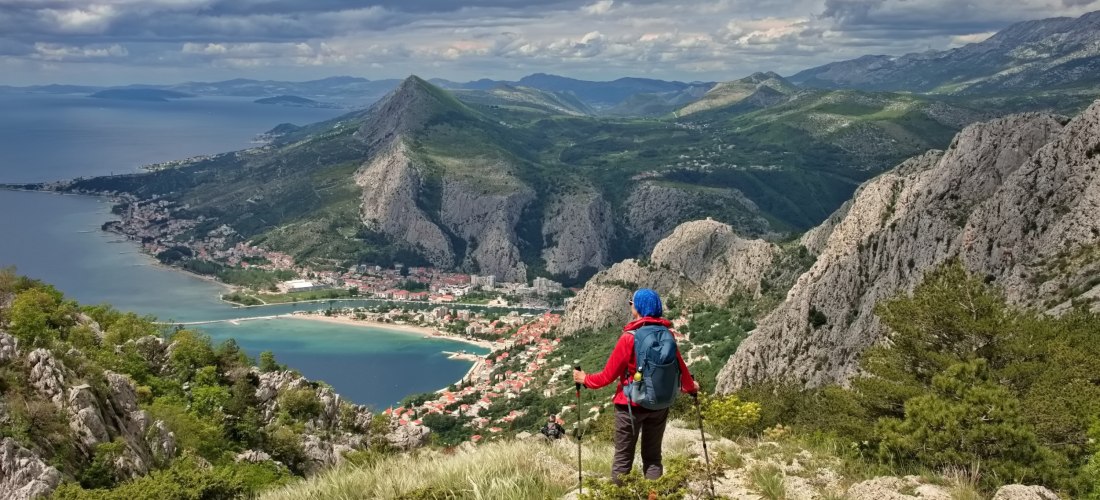Wanderer mit roter Jacke und Rucksack steht auf einem Bergpfad mit Blick auf eine Küstenstadt zwischen Meer und hohen, felsigen Bergen. Der Himmel ist bewölkt, das Panorama weitläufig.