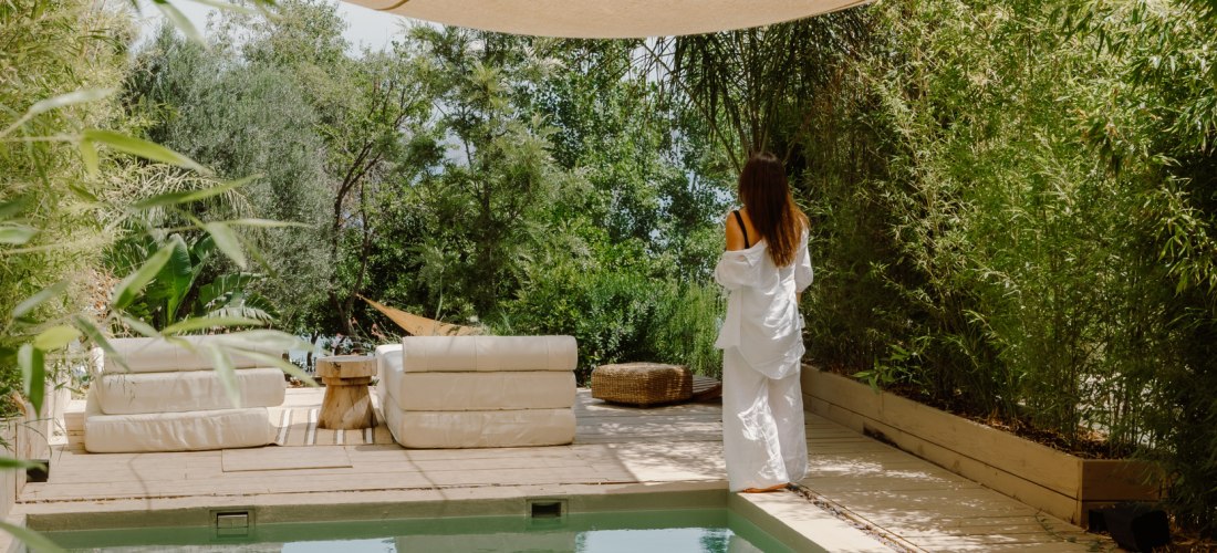 Eine Frau im weißen Bademantel steht auf einer Holzterrasse neben einem kleinen Pool, umgeben von Bambus und mediterraner Vegetation. Über dem Bereich spenden helle Stoffbahnen Schatten, die Atmosphäre ist ruhig und entspannt