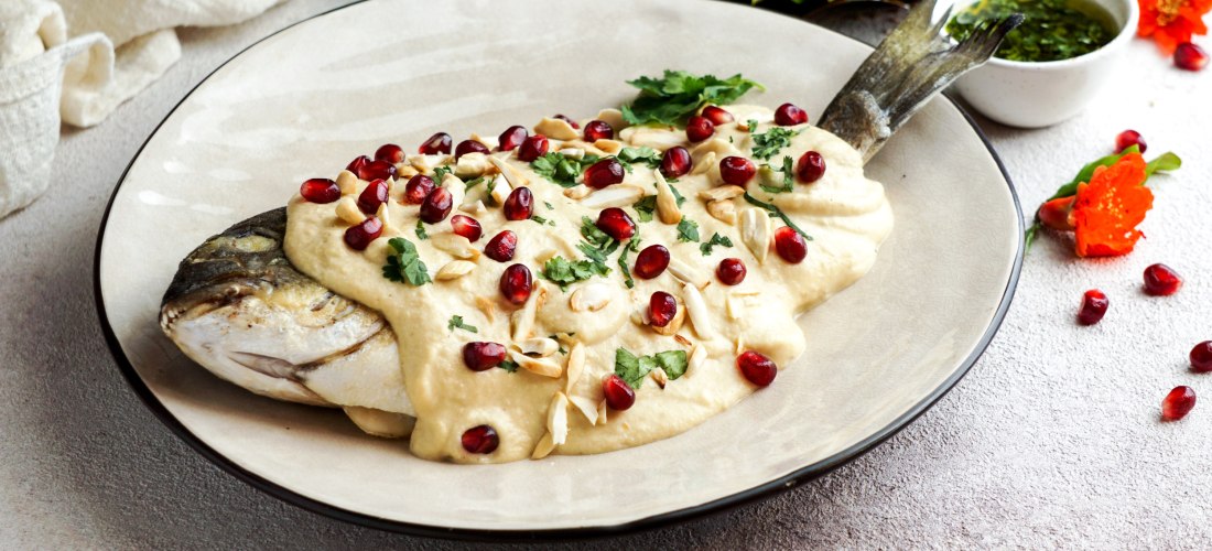 Ein ganzer Fisch auf einem Teller, überzogen mit cremiger Tahini-Sauce, garniert mit Granatapfelkernen, Mandelsplittern und frischen Kräutern. Im Hintergrund Granatapfel, frische Kräuter, eine grüne Sauce und eine orange Blüte.