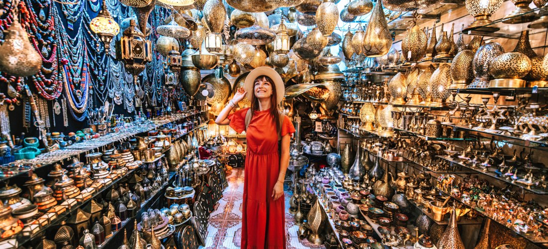 Frau in rotem Kleid und Strohhut steht lächelnd in einem kleinen, überfüllten Souvenir- und Lampengeschäft. Die Wände und Decke sind voller orientalischer Lampen, Laternen, Schmuck und dekorativer Schalen. Das Bild strahlt eine bunte und orientalische Mar