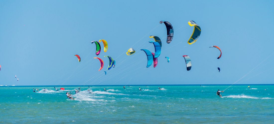 Farbenfrohe Kites segeln am blauen Himmel über dem türkisfarbenen Meer. Zahlreiche Kitesurfer gleiten dynamisch über das Wasser, erzeugen Gischt und nutzen den kräftigen Wind. Die Szene vermittelt Energie, Freiheit und Sportbegeisterung an einem sonnigen 