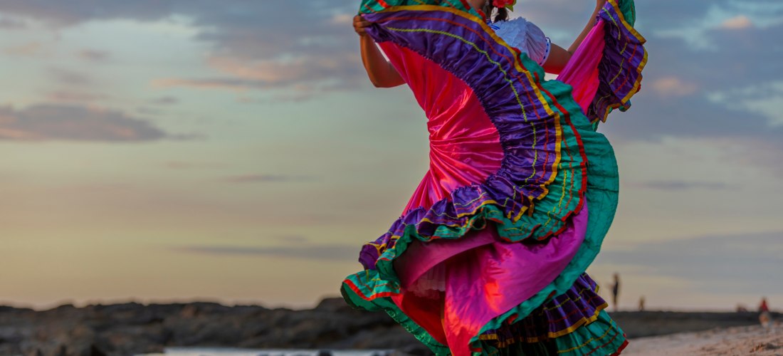 Frau in einem traditionellen, farbenfrohen Kleid tanzt folkloristisch am Strand bei Sonnenuntergang