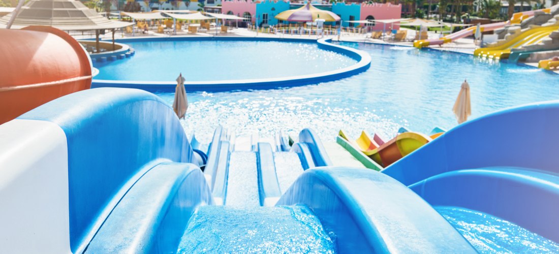 Blick von oben auf mehrere blaue Wasserrutschen, die in einen großen, sonnigen Pool führen. Rund um das Becken stehen Sonnenliegen und bunte Sonnenschirme. Im Hintergrund sind weitere Rutschen in Gelb, Orange und Rot sowie Palmen und bunte Gebäude zu sehe