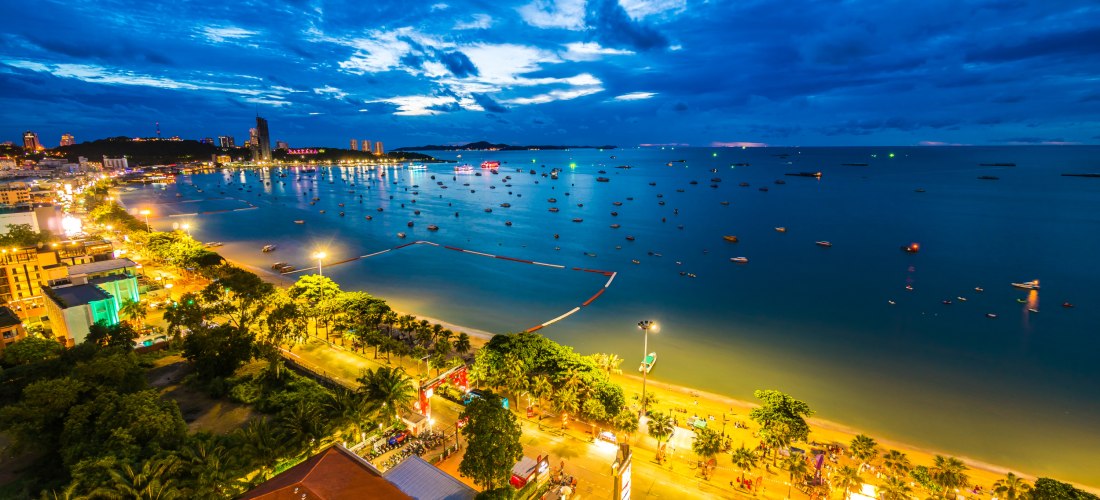 Blick über Pattaya bei Nacht: Die Küste ist hell erleuchtet, zahlreiche Boote liegen in der Bucht. Entlang der Strandpromenade reihen sich Hotels, Palmen und Straßenlichter. Der Himmel ist teils bewölkt, aber noch blau