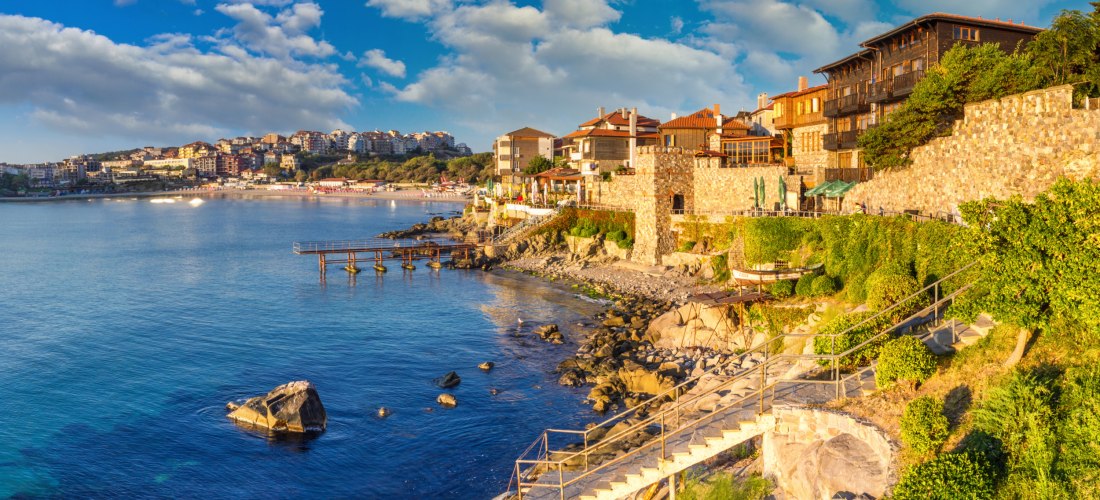 Sonnenuntergang über der Altstadt von Sozopol in Bulgarien. Historische Steinhäuser mit hölzernen Balkonen säumen die felsige Küste, während im Hintergrund moderne Gebäude die Landschaft prägen. Die Treppen führen hinunter zur felsigen Uferlinie, das ruhi