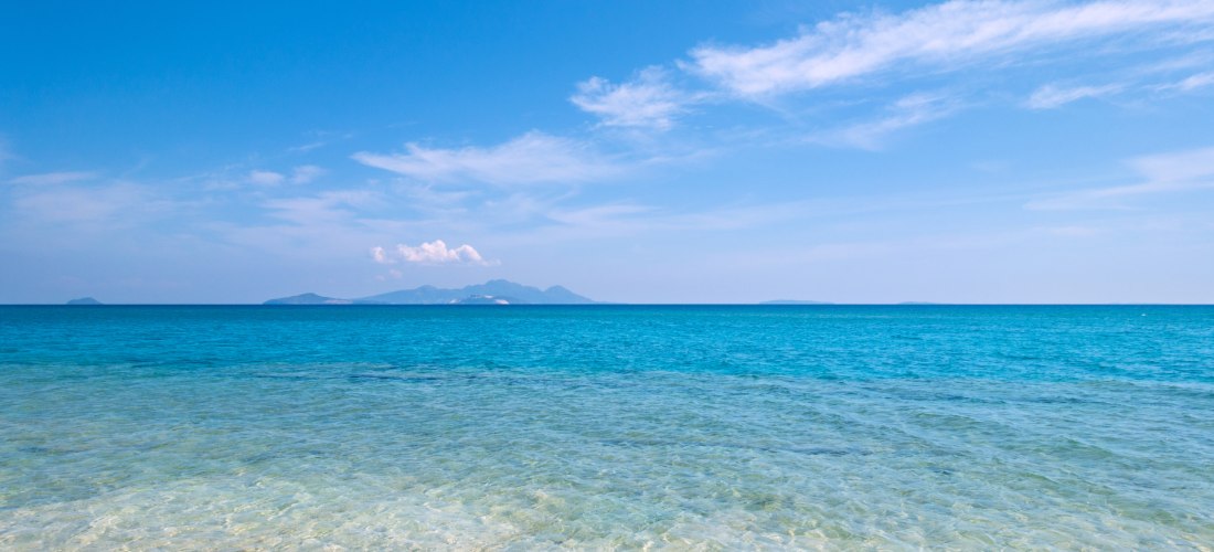 Klares türkisfarbenes Meer mit flachem Wasser am Strand und Blick auf entfernte Inseln unter blauem Himmel mit leichten Wolken