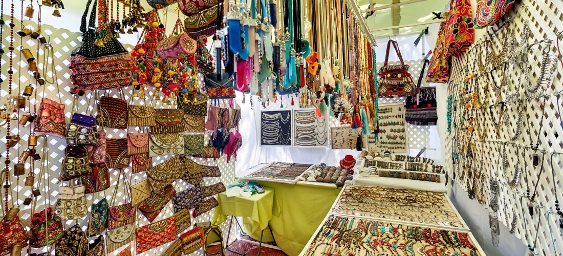 Hippiemarkt auf Ibiza: Ein Stand voller bunter Accessoires. An den Wänden hängen verzierte Handtaschen und Beutel mit Stickereien, während Tische mit Schmuck wie Ketten, Armbändern und Ringen bedeckt sind. Über dem Stand hängen viele Halsketten und dekora