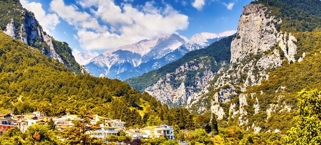 Griechisches Bergdorf am Fuße des Olymps, eingebettet in grüne Wälder mit majestätischen Berggipfeln im Hintergrund.