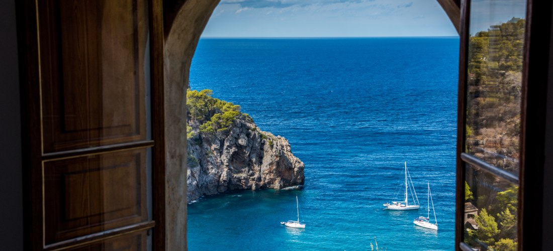 Atemberaubender Blick aus einem offenen Fenster auf das blaue Meer, Klippen und Segelboote bei schönem Wetter auf Mallorca