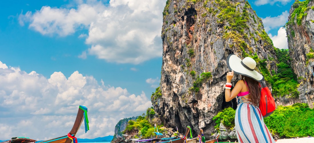 Frau mit Sonnenhut und bunter Sommerkleidung steht am Strand und blickt auf traditionelle Langboote und eine steile Felsformation. Türkisfarbenes Meer, blauer Himmel mit Wolken