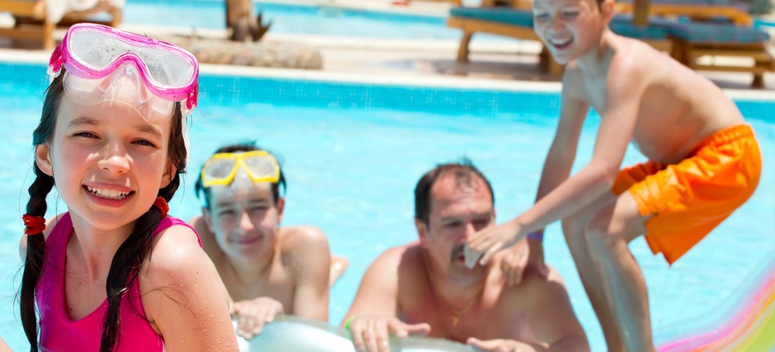 Mehrere Kinder und ein Vater spielen fröhlich in einem Hotelpool. Ein Mädchen mit rosa Badeanzug und Schwimmbrille lächelt in die Kamera, während ein Junge auf einer Luftmatratze steht. Im Hintergrund liegen weitere Gäste auf Sonnenliegen