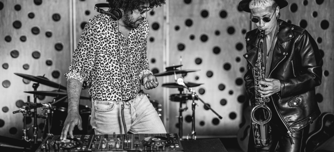 Schwarz-weiß-Foto eines Musikduos: Links ein DJ mit Leopardenhemd an einem Mischpult, rechts ein Saxophonist mit Sonnenbrille, Hut und Lederjacke beim Spielen des Saxophons. Im Hintergrund Schlagzeug-Setup.