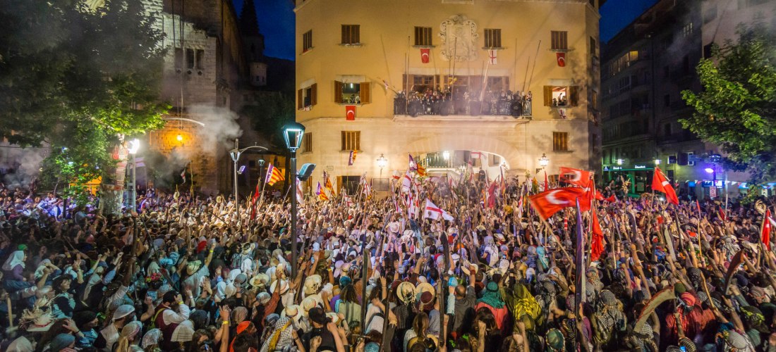 Große Menschenmenge auf einem Platz vor dem Rathaus von Sóller auf Mallorca bei Nacht. Viele Menschen tragen Kopftücher und Hüte, schwenken Fahnen und Stöcke. Auf dem Balkon des historischen Gebäudes stehen Menschen in festlicher Kleidung. Das Fest ist Te