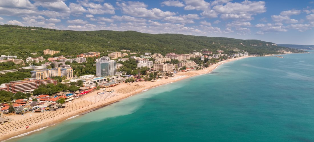 Luftaufnahme vom Goldstrand in Bulgarien. Entlang der Küste reihen sich zahlreiche Hotels und Ferienanlagen aneinander, vor einer üppig grünen Hügellandschaft. Der feinsandige Strand ist mit Liegen und Sonnenschirmen ausgestattet, das türkisfarbene Wasser