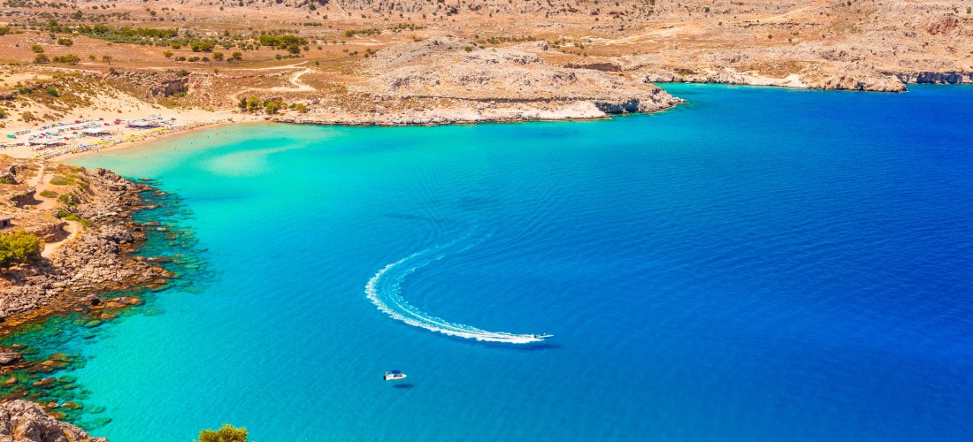  Das Bild zeigt eine weite, halbmondförmige Bucht auf Rhodos, nahe Lindos, mit glasklarem türkisfarbenem Wasser.    Am linken Rand des Strands befinden sich zahlreiche Sonnenschirme und Liegen.    Zwei Boote bewegen sich auf dem Wasser – eines davon hinte