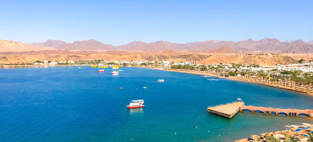 Blick auf eine weitläufige Badebucht mit türkisblauem Wasser, gesäumt von Palmen und Sonnenschirmen. Am Ufer führt eine Promenade mit Steg ins Meer. Mehrere Ausflugsboote und Yachten liegen auf dem Wasser. Im Hintergrund sind Wüstenberge und eine flache S
