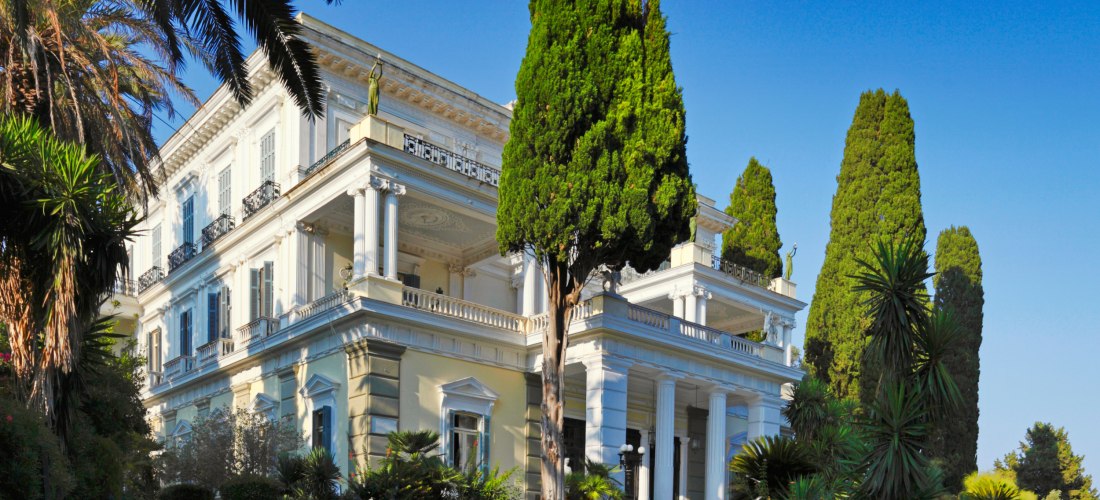 Der Achilleion-Palast auf Korfu, ein weißes klassizistisches Gebäude mit Säulen, Balkonen und Statuen auf dem Dach. Umgeben von hohen Zypressen, Palmen und Blumenbeeten vermittelt der Palast ein majestätisches Flair unter strahlend blauem Himmel.
