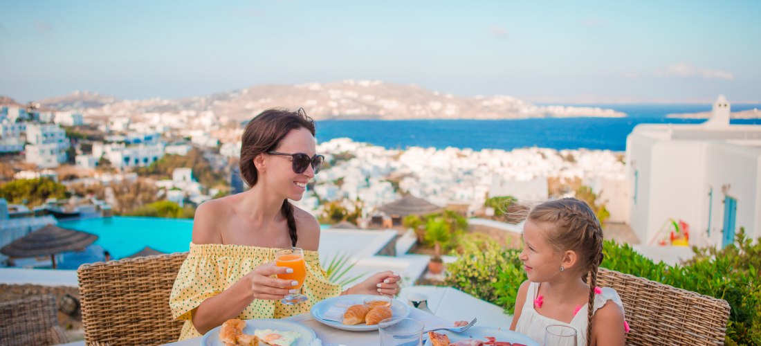 Eine Frau und ein kleines Mädchen frühstücken an einem Tisch im Freien mit Blick auf eine Küstenstadt mit weißen Häusern und das blaue Meer. Die Frau trägt ein gelbes Sommerkleid und hält ein Glas Orangensaft, beide lächeln sich an. Auf dem Tisch liegen T