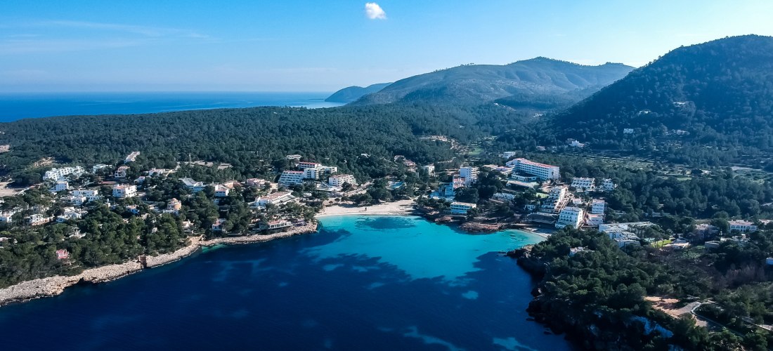 Luftaufnahme eines kleinen Ferienortes im Norden Ibizas. Der Ort liegt zwischen bewaldeten Hügeln am türkisblauen Meer mit Stränden, Hotels und Ferienhäusern.