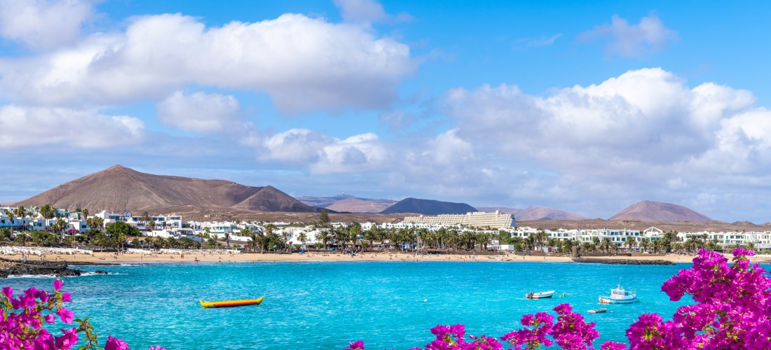 Panoramablick auf Costa Teguise auf Lanzarote mit hellem Sandstrand, türkisblauem Meer, weißen Häusern und Bergen im Hintergrund.