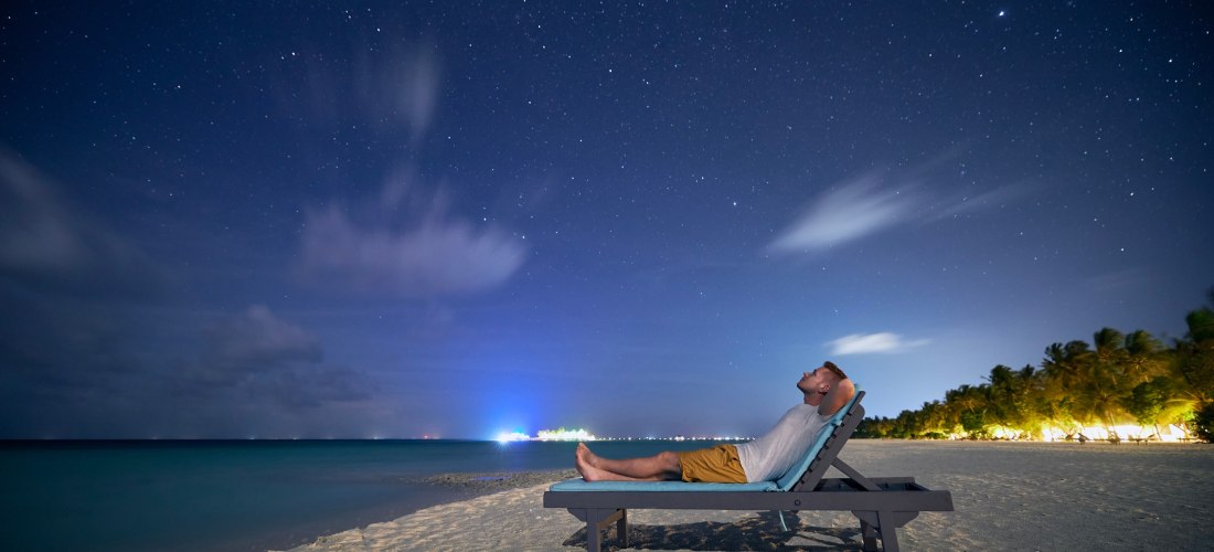  Ein Mann liegt entspannt auf einer Strandliege und blickt in den funkelnden Sternenhimmel.              Der ruhige Ozean, die sanft beleuchteten Palmen und die friedliche Nachtstimmung machen diesen Moment magisch und unvergesslich.
