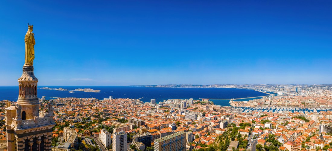 Weitwinkliges Stadtpanorama einer südfranzösischen Küstenmetropole. Links im     Vordergrund ragt ein Turm mit einer goldenen Marienstatue auf. Darunter breitet     sich die Stadt mit vielen orangefarbenen Ziegeldächern und modernen Hochhäusern aus.     M