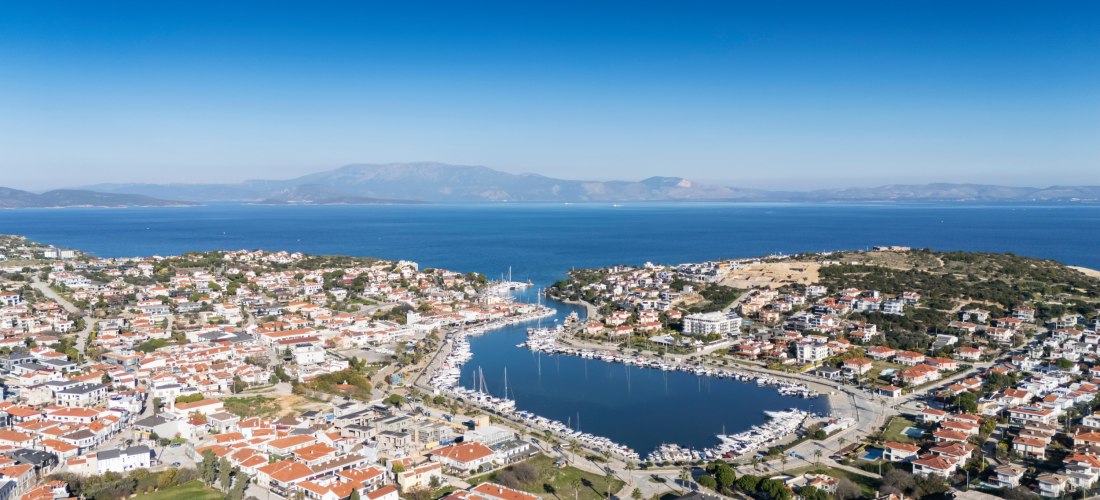 Panoramablick auf das Küstenstädtchen Çeşme an der türkischen Ägäis mit rotdachigen Häusern, einem Yachthafen in der Mitte und dem tiefblauen Meer im Hintergrund. Am Horizont sind sanfte Hügel und griechische Inseln zu sehen.
