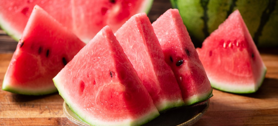  Das Bild zeigt mehrere dreieckig geschnittene Stücke Wassermelone, die auf einem kleinen Teller angerichtet sind.   Das saftige rote Fruchtfleisch ist mit wenigen dunklen Kernen durchsetzt und wird von einer dünnen, grünen Schale umrandet.   Im Hintergru