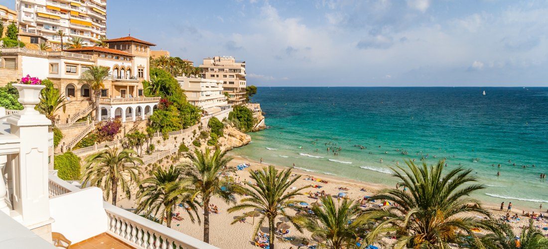 Belebter Stadtstrand mit vielen Badegästen, Palmen und Sonnenschirmen. Das türkisfarbene Meer glitzert in der Sonne, während sich Menschen im Wasser und am Strand entspannen. Im Hintergrund ragen moderne Apartmenthäuser und historische Villen über dem Str