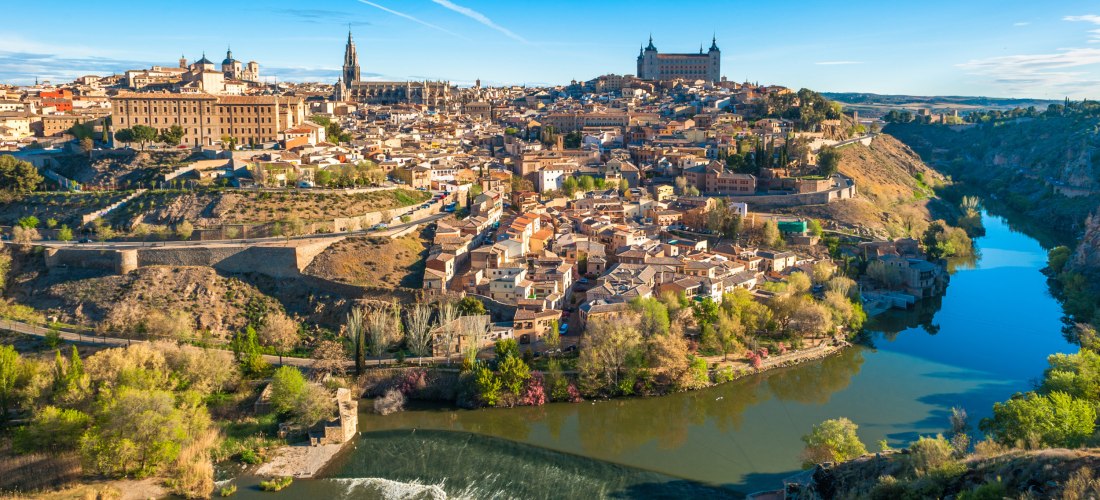 Panoramablick auf die Altstadt von Toledo, Spanien, umgeben vom Fluss Tajo. Gut sichtbar sind die Kathedrale von Toledo und der Alcázar. Die Stadt liegt malerisch auf einem Hügel, umgeben von grüner Landschaft.