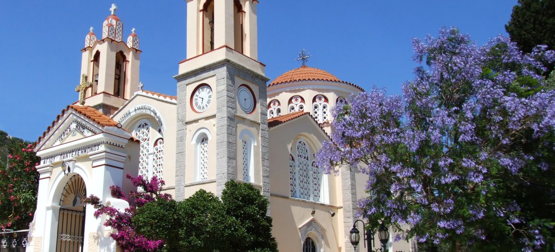 Griechisch-orthodoxe Kirche mit zwei Glockentürmen und einer Kuppel, verziert mit weißen Ornamenten und Kreuzen. Vor dem Gebäude stehen blühende Bougainvillea und ein Baum mit violetten Blüten. Die Fassade ist hell gestrichen, das Dach mit roten Ziegeln g