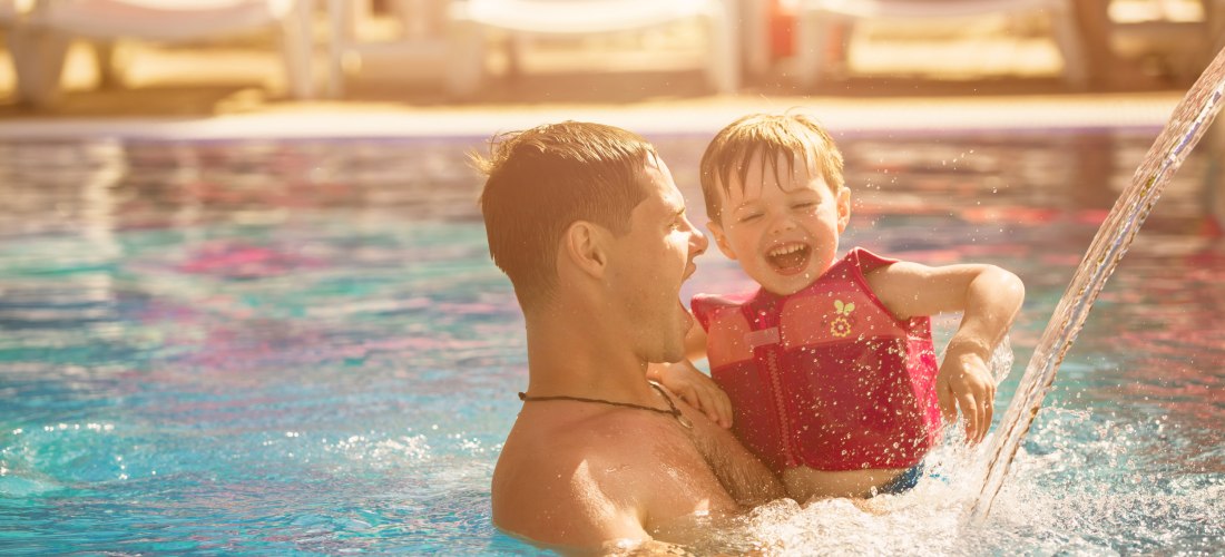 Vater spielt mit kleinem Kind in einem Pool unter freiem Himmel, beide lachen ausgelassen