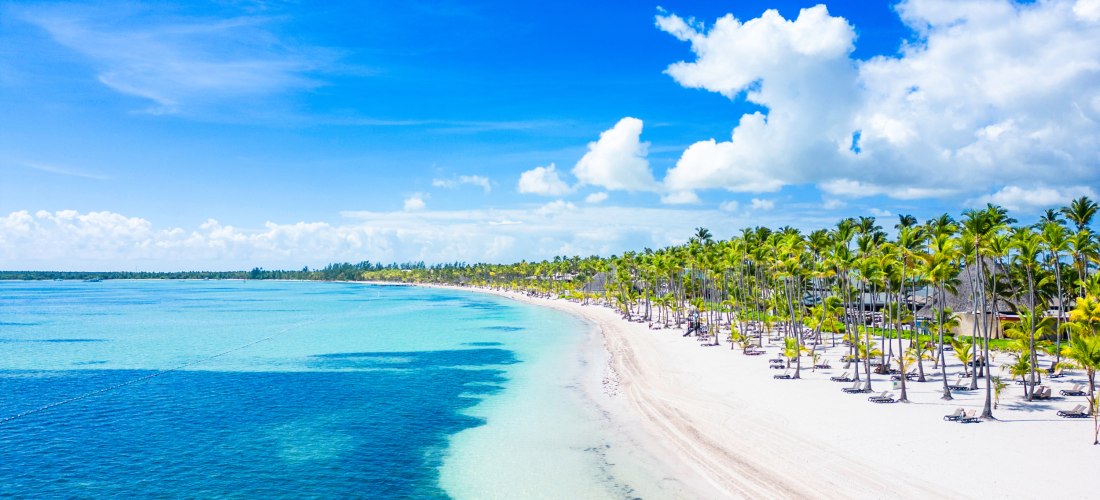 Endloser, weißer Sandstrand in der Dominikanischen Republik, gesäumt von hohen Palmen. Das türkisblaue, glasklare Meer trifft unter strahlend blauem Himmel auf das Ufer. Die Szene vermittelt Ruhe, Karibikfeeling und perfekte Bedingungen für einen entspann