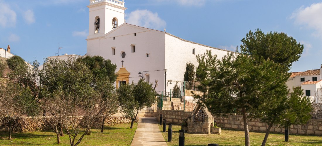 Weiß getünchte Kirche mit Glockenturm in Es Mercadal, Menorca. Ein gepflasterter Weg führt durch eine kleine Grünanlage mit Bäumen zur Kirche.