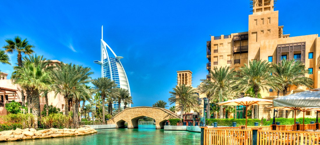 Malereihafter Anblick des Burj Al Arab in Dubai, umgeben von Palmen, traditioneller Architektur und türkisblauem Wasser. Eine kleine Steinbrücke führt über den Kanal, Sonnenschirme und Terrassen laden am Ufer zum Verweilen ein, während der Himmel strahlen