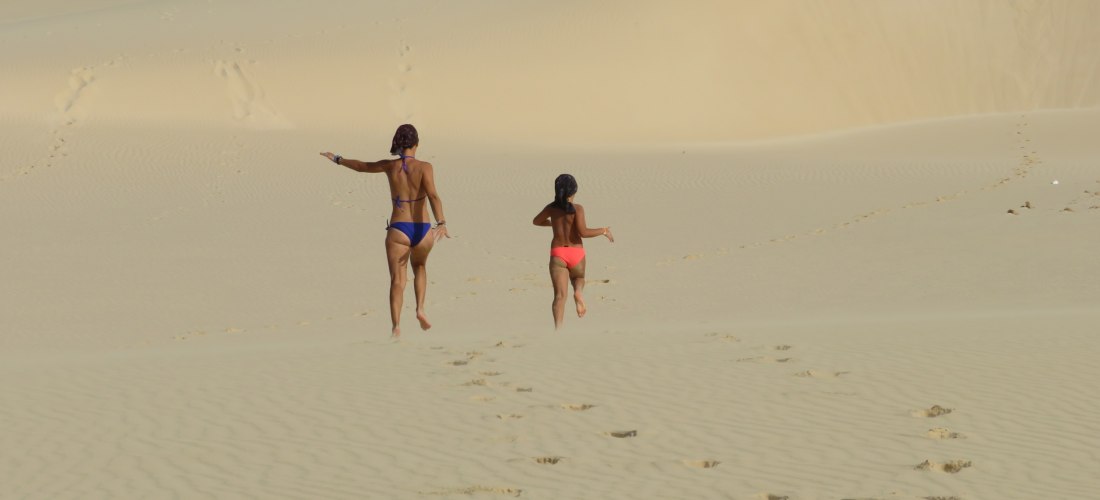 Zwei Personen in Badebekleidung laufen barfuß durch weitläufige Sanddünen. Ihre Fußspuren zeichnen sich im feinen, hellen Sand ab. In der Ferne ist eine weitere Person zu sehen, die eine Düne erklimmt. Die Szenerie wirkt ruhig und endlos